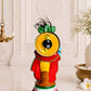 A colorful decorative figurine of a stylized human figure with a large yellow sunflower head, red and yellow clothing, and a black base, displayed on a light marble surface with a hanging brass lamp, white flowers in a vase, and a small white flower on a table runner.