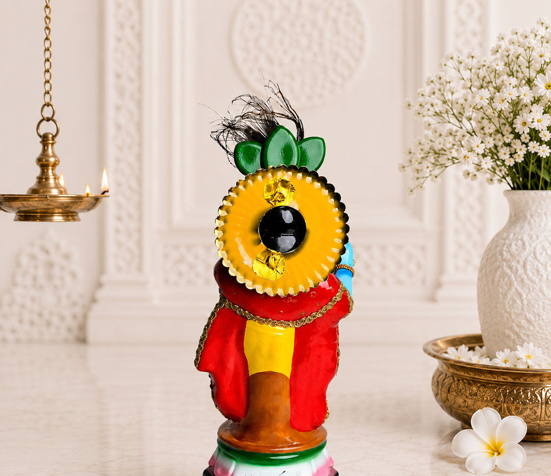 A colorful decorative figurine of a stylized human figure with a large yellow sunflower head, red and yellow clothing, and a black base, displayed on a light marble surface with a hanging brass lamp, white flowers in a vase, and a small white flower on a table runner.