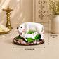 A white cow and calf figurine with green accents on a brown textured base, shown on a beige surface with decorative props in the background and measurement arrows indicating 4.5 inches wide by 3.