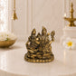 A small gold-toned brass idol of Lord Hanuman and Gauravendra on a decorative pedestal, displayed on a white marble surface with a softly blurred background featuring a brass lamp, flowers, and a hanging bell.