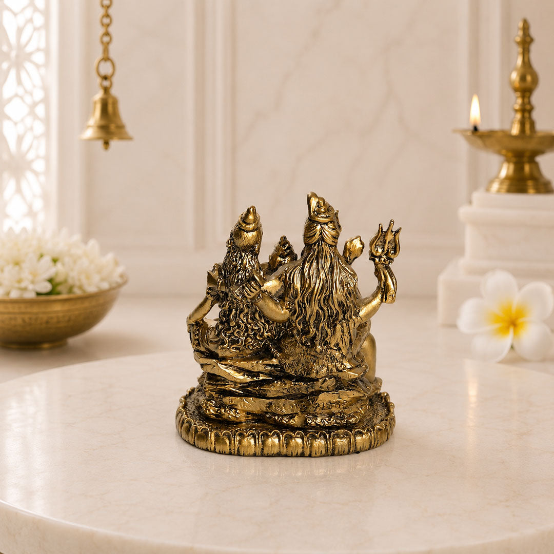 A small gold-toned brass idol of Lord Hanuman and Gauravendra on a decorative pedestal, displayed on a white marble surface with a softly blurred background featuring a brass lamp, flowers, and a hanging bell.